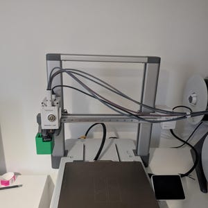 May include: A 3D printer with a silver frame, black and grey components, and a green accent. The printer has a rectangular build plate with a geometric design. Cables and wires are visible, along with a spool of filament.