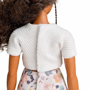 May include: A doll wearing a white short-sleeved top and a floral print skirt. The top has a textured, ribbed design. The skirt features a pattern of pink, blue, and yellow flowers on a white background. The doll has brown hair.