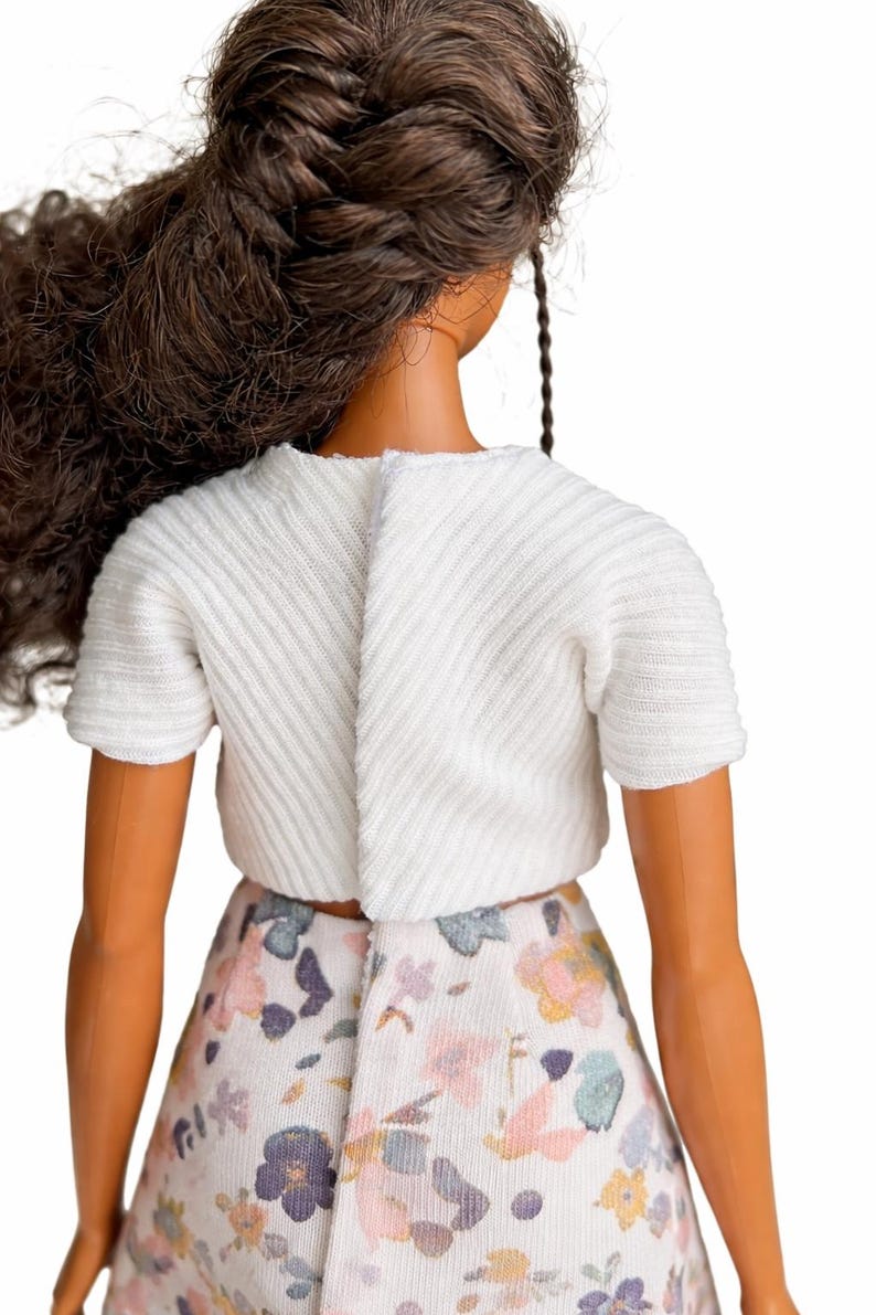 May include: A doll wearing a white short-sleeved top and a floral print skirt. The top has a textured, ribbed design. The skirt features a pattern of pink, blue, and yellow flowers on a white background. The doll has brown hair.