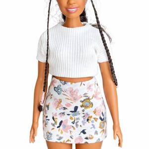 May include: A doll with dark hair styled in braids, wearing a white ribbed crop top and a floral print mini skirt. The skirt features a pattern of pink, blue, and yellow flowers on a white background. The doll has a smiling expression.