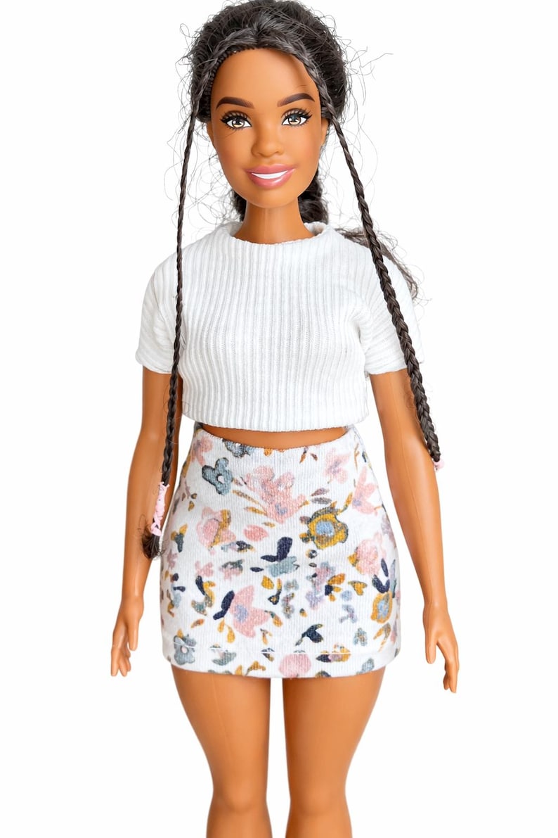 May include: A doll with dark hair styled in braids, wearing a white ribbed crop top and a floral print mini skirt. The skirt features a pattern of pink, blue, and yellow flowers on a white background. The doll has a smiling expression.
