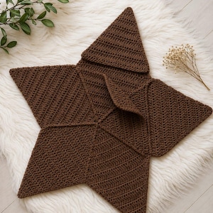 May include: A dark brown star-shaped blanket crafted with a textured crochet stitch pattern. The item features five distinct points and a folded section in the center, showcasing a consistent yarn construction throughout the entire piece.