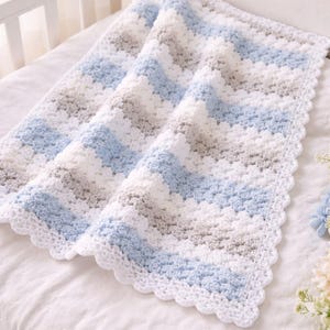 May include: A handmade baby blanket in a crib. The blanket has a chevron pattern of light blue, white, and grey stripes. The edges are finished with a white, scalloped border. The blanket is approximately 91 cm by 102 cm.