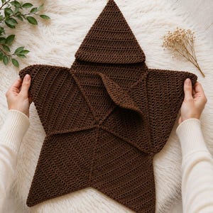May include: A dark brown star-shaped crochet blanket featuring a textured stitch pattern. The item has five distinct points and a folded center section, crafted from yarn with a consistent dark brown color throughout the entire piece.