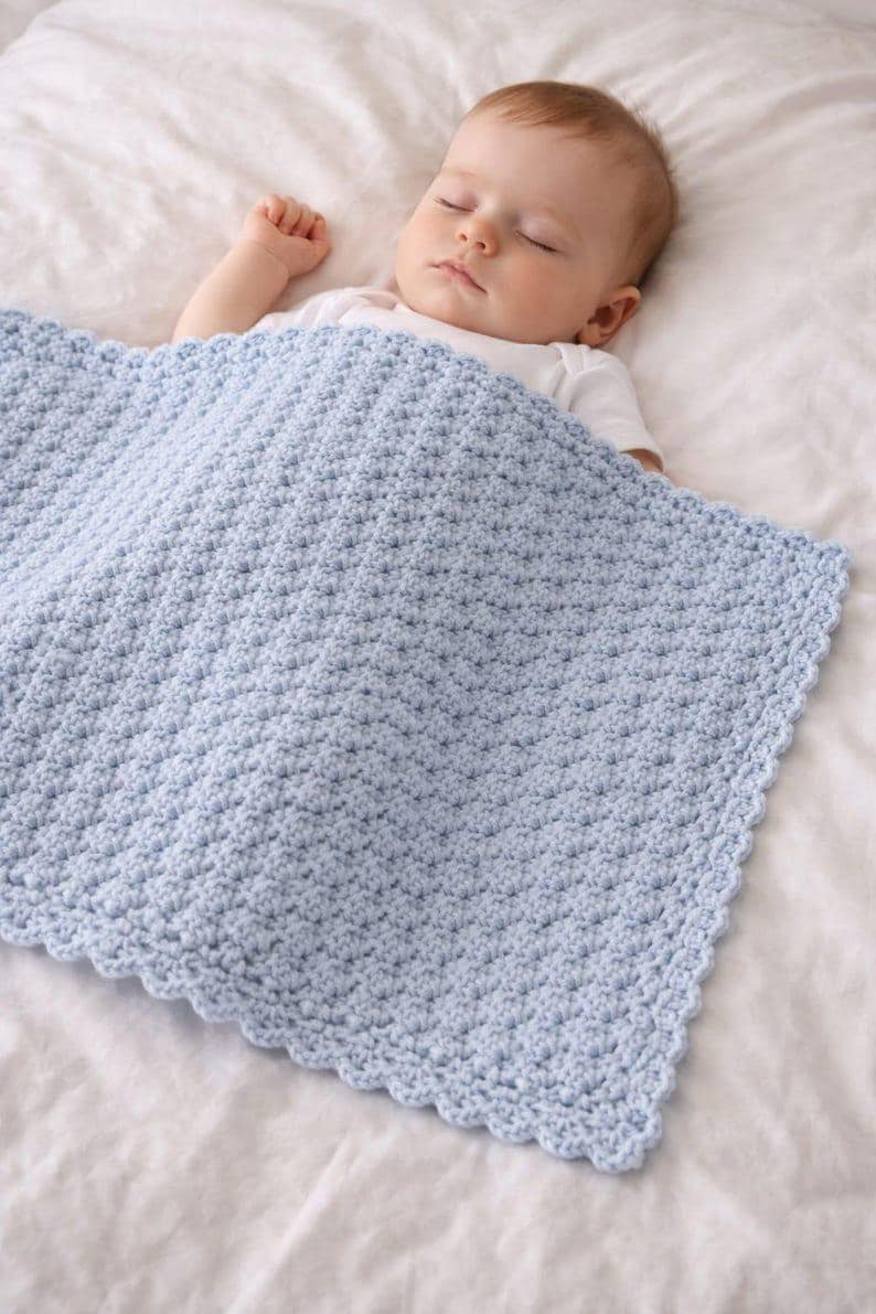 K&ouml;nnte beinhalten: Eine hellblaue Babydecke mit einem strukturierten, sich wiederholenden Stichmuster und einem gewellten Rand. Die Decke ist rechteckig geformt und weist eine durchgehend konsistente und dichte H&auml;kelkonstruktion auf der gesamten Oberfl&auml;che auf.