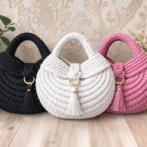 May include: Three handmade crochet handbags in black, white, and pink. Each bag features a rounded shape, a top handle, and a gold-colored clasp with a matching tassel. The bags are displayed on a wooden surface against a floral patterned background.