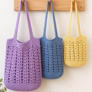 May include: Three crochet tote bags in purple, blue, and yellow, hanging from a wooden rack. The bags feature an openwork stitch pattern with solid bases and handles. The bags are labelled as LARGE, MEDIUM, and SMALL to indicate their sizes.