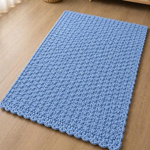K&ouml;nnte beinhalten: Ein rechteckiger hellblauer Teppich mit einer dicken, groben Strickstruktur, einem sich wiederholenden Noppenmuster und einem gewellten Randabschluss. Der Teppich ist aus weichem Garn gefertigt und weist eine gleichm&auml;&szlig;ige Farbe auf seiner Oberfl&auml;che auf.