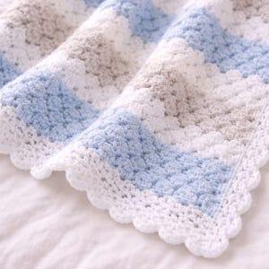K&ouml;nnte beinhalten: Eine geh&auml;kelte Decke mit einem sich wiederholenden Muster aus horizontalen Streifen in Hellblau, Wei&szlig; und sanftem Beige. Die Decke ist mit einem strukturierten Stich gefertigt und mit einem dekorativen Muschelrand in wei&szlig;em Garn versehen.