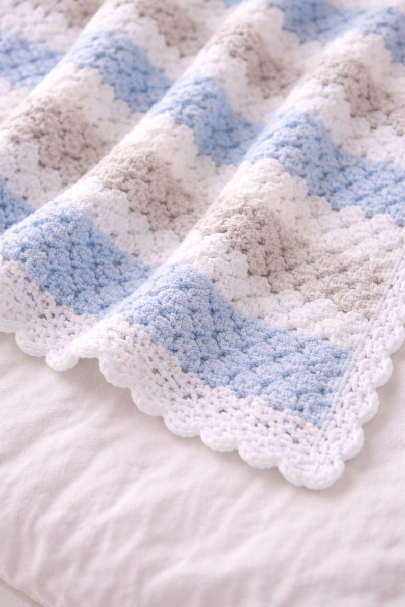 K&ouml;nnte beinhalten: Eine geh&auml;kelte Decke mit einem sich wiederholenden Muster aus horizontalen Streifen in Hellblau, Wei&szlig; und sanftem Beige. Die Decke ist mit einem strukturierten Stich gefertigt und mit einem dekorativen Muschelrand in wei&szlig;em Garn versehen.