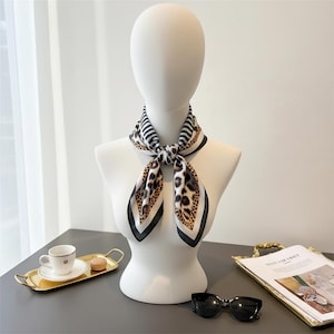 Può includere: Una sciarpa di seta con un motivo a righe bianche e nere e stampa leopardata è drappeggiata attorno al collo di un manichino. La sciarpa ha un bordo nero. Un vassoio dorato con una tazza e un piattino e occhiali da sole sono sul tavolo.