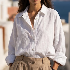 May include: A white linen button-down shirt with long sleeves and a classic collar. The shirt is partially unbuttoned, revealing the neckline. The sleeves are rolled up. Paired with tan-colored high-waisted pants. The outfit is simple and elegant.
