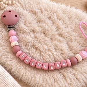 May include: A dummy clip featuring a mauve silicone clip, a string of round and ribbed beads in shades of pink and mauve, and square beads spelling out the word PRINCESS in white letters, finished with a pink cord loop at the end.