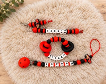 May include: A set of three baby accessories featuring red and black silicone beads and white letter beads spelling the name KIERAN. The set includes a pacifier clip, a teething ring, and a keychain, each accented with a red race car charm.