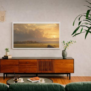Op de afbeelding: Een woonkamer met een ingelijst landschapsschilderij boven een houten kast. Een groene bank, een ronde salontafel en een grote plant in pot zijn aanwezig. Een witte luidspreker en decoratieve wandkleden zijn ook zichtbaar.