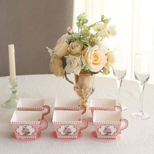 May include: Six pink paper teacups with matching saucers are arranged on a white tablecloth. Each teacup features a floral design and a scalloped edge. A bouquet of cream-colored roses sits in a decorative vase, with champagne flutes in the background.