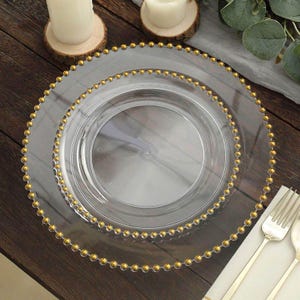 Op de afbeelding: Twee heldere glazen onderborden met een gouden kralenrand. De borden staan op een donker houten oppervlak, met een vork en lepel rechts. De borden zijn ontworpen voor decoratief gebruik.