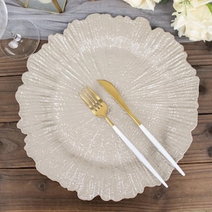 May include: A round, beige glass charger plate with a textured, radiating sunburst pattern. A gold-toned metal fork and a matching gold-toned metal knife, both featuring sleek white handles, are placed diagonally across the center of the plate.