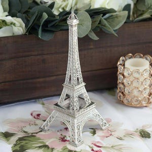 Peut inclure: Une réplique argentée de la Tour Eiffel, un objet décoratif, est posée sur une surface à motifs floraux. La tour est finement détaillée avec un treillis ajouré et est une version miniature du monument emblématique. Un bougeoir avec des cristaux est en arrière-plan.