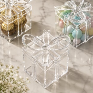 Könnte beinhalten: Drei quadratische Geschenkboxen aus klarem Acryl mit abnehmbaren Deckeln, die oben mit einer geformten Schleife versehen sind. Eine Box ist leer, eine ist mit goldverpackten Pralinen und eine mit bunten, runden Pastellbonbons gefüllt.