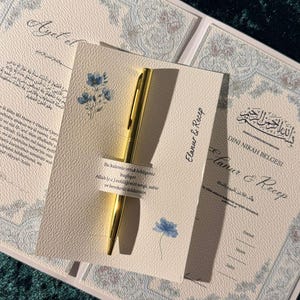 Könnte beinhalten: Eine cremefarbene Karte mit einem befestigten goldenen Stift, verziert mit blauen Blumenillustrationen und türkischem Text. Daneben liegt ein passendes Dokument mit dem Titel Dini Nikah Belgesi, den Namen Elanur & Recep sowie arabischer Schrift.