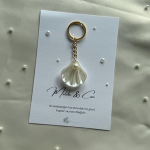 Könnte beinhalten: Ein muschelförmiger Schlüsselanhänger mit Perlenakzent, befestigt an einem goldfarbenen Ring. Der Schlüsselanhänger hängt an einer weißen Karte mit den Namen "Melda & Can" und einer türkischen Nachricht. Streu-Perlen sind im Hintergrund.