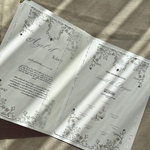 Peut inclure: Un document rectangulaire blanc ouvert avec des coins en métal doré. La page de gauche présente le texte Ayet el Kursi en arabe et en anglais. La page de droite est intitulée Dini Nikah Belgesi avec les noms Saudet & Dajan imprimés dessous.