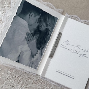 Könnte beinhalten: Eine weiße Hochzeitsmappe mit einem Schwarz-Weiß-Foto eines Paares auf der linken Seite und deutschem Text auf der rechten Seite. Ein silberner Stift ist in der Mitte befestigt. Die Mappe ist an den Rändern mit weißer Spitze verziert.