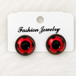 Puede incluir: Un par de pendientes redondos con fondo rojo brillante y lunares negros, engastados en un marco de metal color bronce oscuro. Los pendientes están sujetos a una tarjeta expositora rectangular blanca con el texto Fashion Jewelry impreso en negro.