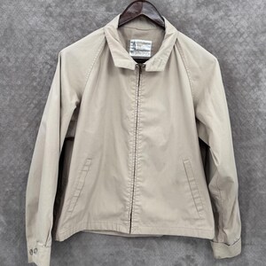 May include: A tan jacket with a full zipper closure, two front pockets, and a collared neckline. The jacket is hanging on a wooden hanger. The label on the inside of the collar is visible.