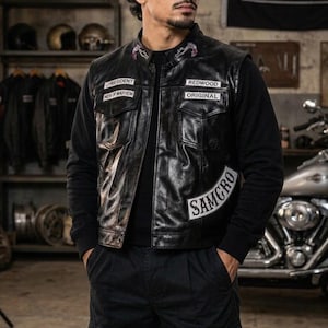 Könnte beinhalten: Eine schwarze Leder-Motorradweste mit weißen gestickten Aufnähern, die UPRESIDENT, MEN OF MAYHEM, REDWOOD, ORIGINAL und SAMCRO zeigen. Die Weste hat zwei Brusttaschen, einen Kragen mit kleinen Stickereien und wird über einem schwarzen Langarmshirt getragen.