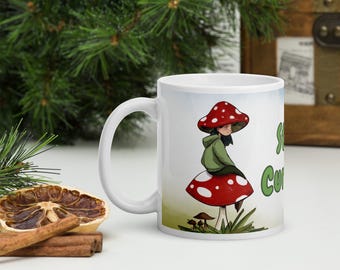 Puede incluir: Una taza de cerámica blanca con una ilustración gráfica de una persona con sudadera verde sentada sobre una seta grande roja con puntos blancos. El texto "STAY CURIOUS" está impreso en letras verdes en el lateral de la taza junto al dibujo.