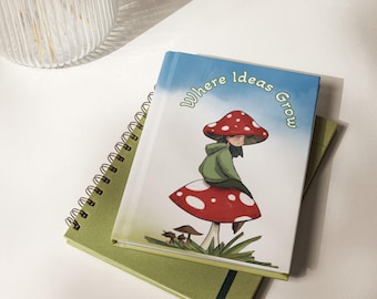 Puede incluir: Un diario de tapa dura con cubierta blanca que muestra un hongo rojo con puntos blancos, una persona con sudadera verde sentada sobre él y el texto Where Ideas Grow, sobre un cuaderno de espiral verde con una banda elástica verde oscuro.