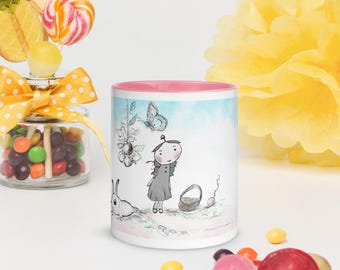 Puede incluir: Taza de cerámica blanca con interior rosa, con una ilustración caprichosa de una niña, un conejo y una mariposa. El diseño tiene un fondo de acuarela con un degradado azul claro y rosa. Caramelos y una flor amarilla en el fondo.
