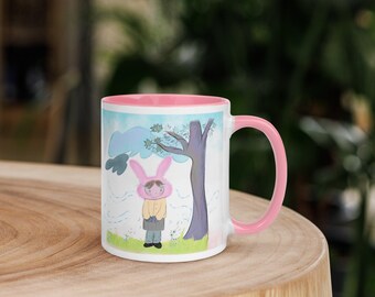 Puede incluir: Taza de cerámica blanca con interior y asa rosas. La taza presenta una ilustración caprichosa de una persona con un disfraz de conejito rosa de pie cerca de un árbol. El fondo incluye un cielo azul y una zona de césped.