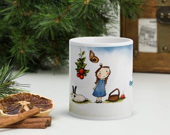 Puede incluir: Una taza de cerámica blanca con una ilustración de una niña con trenzas castañas y vestido azul, acompañada por un conejo blanco, una cesta marrón, una manzana roja, una mariposa y una rama con bayas rojas sobre un campo verde.