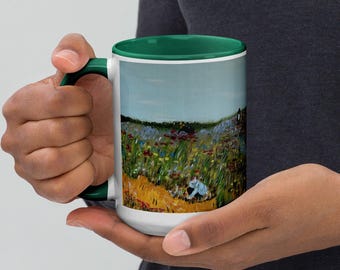Puede incluir: Una taza de cerámica blanca con interior y asa de color verde oscuro, que presenta una reproducción impresa de un paisaje con un campo de flores silvestres, hierbas altas de color verde y una pequeña figura con camisa azul claro.