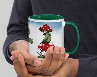 Puede incluir: Una taza de cerámica blanca con interior y asa de color verde oscuro, que presenta una ilustración gráfica de una persona con un sombrero de seta roja sentada sobre una seta grande de color rojo con puntos blancos.