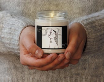 Puede incluir: Una vela encendida en un tarro de cristal transparente. El tarro tiene una etiqueta negra con una ilustración en blanco y negro de una mujer con los ojos cerrados y el texto "She stops existing... and suddenly nothing binds her."