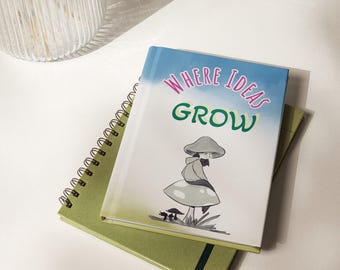 Puede incluir: Un diario de tapa dura con un degradado azul y blanco, con el texto "WHERE IDEAS GROW" en rosa y verde, y una ilustración de una persona sentada sobre una seta. El diario descansa sobre un cuaderno de espiral de color verde.
