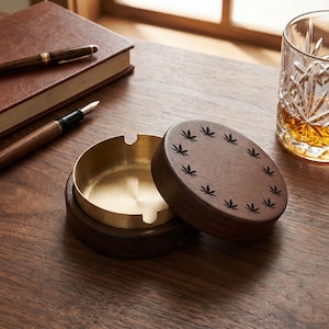 May include: A wooden ashtray with a brass interior and a lid decorated with cannabis leaf designs. The ashtray sits on a wooden surface, next to a glass of amber liquid, a notebook, and pens.