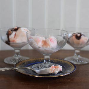 Puede incluir: Tres cuencos de postre de vidrio transparente llenos de helado rosa cubierto con salsa de chocolate. Una cuchara de plata con una bola de helado rosa encima se encuentra sobre un plato decorativo azul y dorado.