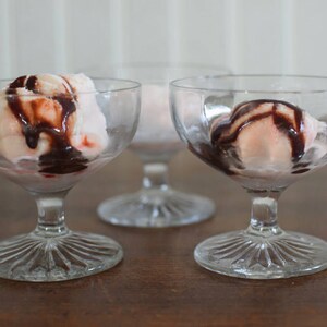 Puede incluir: Tres copas de postre de vidrio transparente con helado blanco y rosa cubierto con salsa de chocolate.