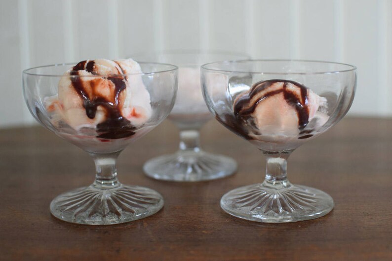 Puede incluir: Tres copas de postre de vidrio transparente con helado blanco y rosa cubierto con salsa de chocolate.