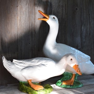 May include: Two white ceramic duck figurines with glossy finishes. One duck stands tall with its orange beak open, while the other is positioned lower with its head tilted downward. Both ducks feature bright orange feet and rest on green bases.