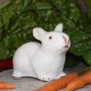 May include: A white ceramic rabbit figurine with pink-toned eyes and nose details. The rabbit is positioned in a seated posture with upright ears, featuring a smooth, glossy finish and visible molded facial features including whiskers and a mouth.