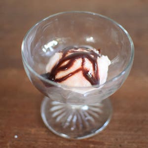 Puede incluir: Un bol de cristal transparente con una bola de helado de vainilla cubierta con salsa de chocolate.