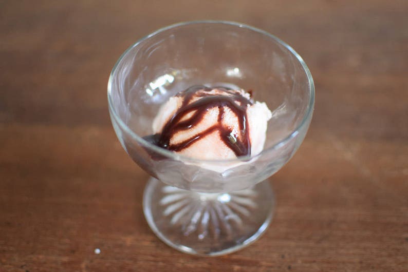 Puede incluir: Un bol de cristal transparente con una bola de helado de vainilla cubierta con salsa de chocolate.