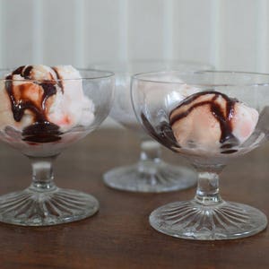 Puede incluir: Tres copas de postre de vidrio transparente con helado rosa cubierto con salsa de chocolate.