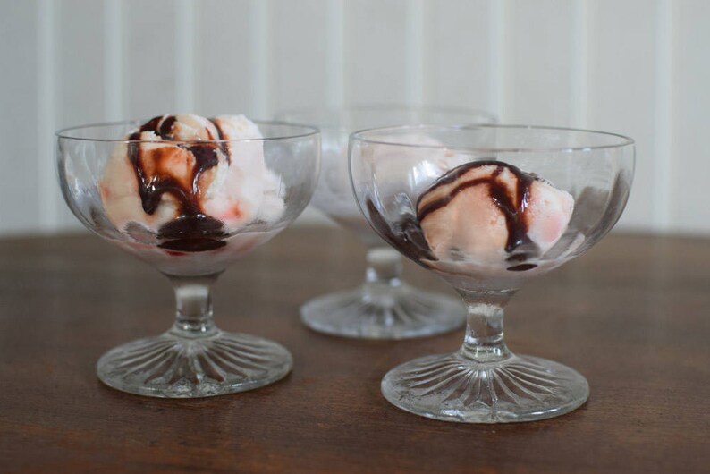 Puede incluir: Tres copas de postre de vidrio transparente con helado rosa cubierto con salsa de chocolate.